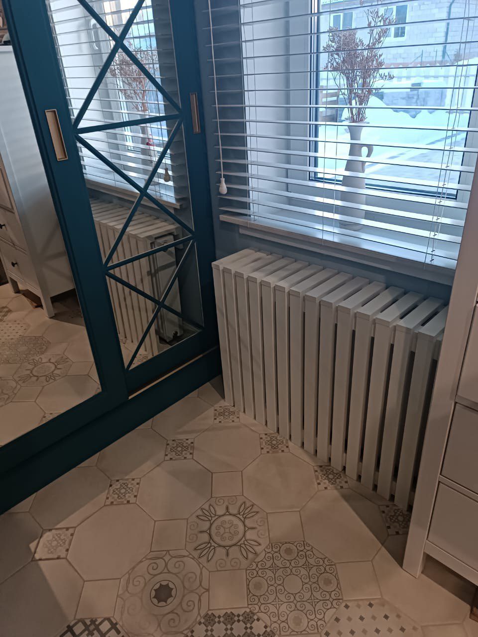 Белый вертикальный трубчатый радиатор отопления и синяя межкомнатная дверь с остеклением и ромбовидной раскладкой на фоне узорчатой плитки. Пример отделки лоджии в [dynamic id="city" column="prepositional"]